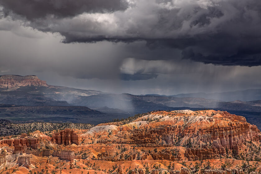 Rain falls from dark clouds above a red-orange landscape of hoodooes and coniferous trees.