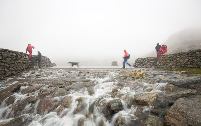 A man walking his dog over a weir at Levers Water reservoir in the Lake District.